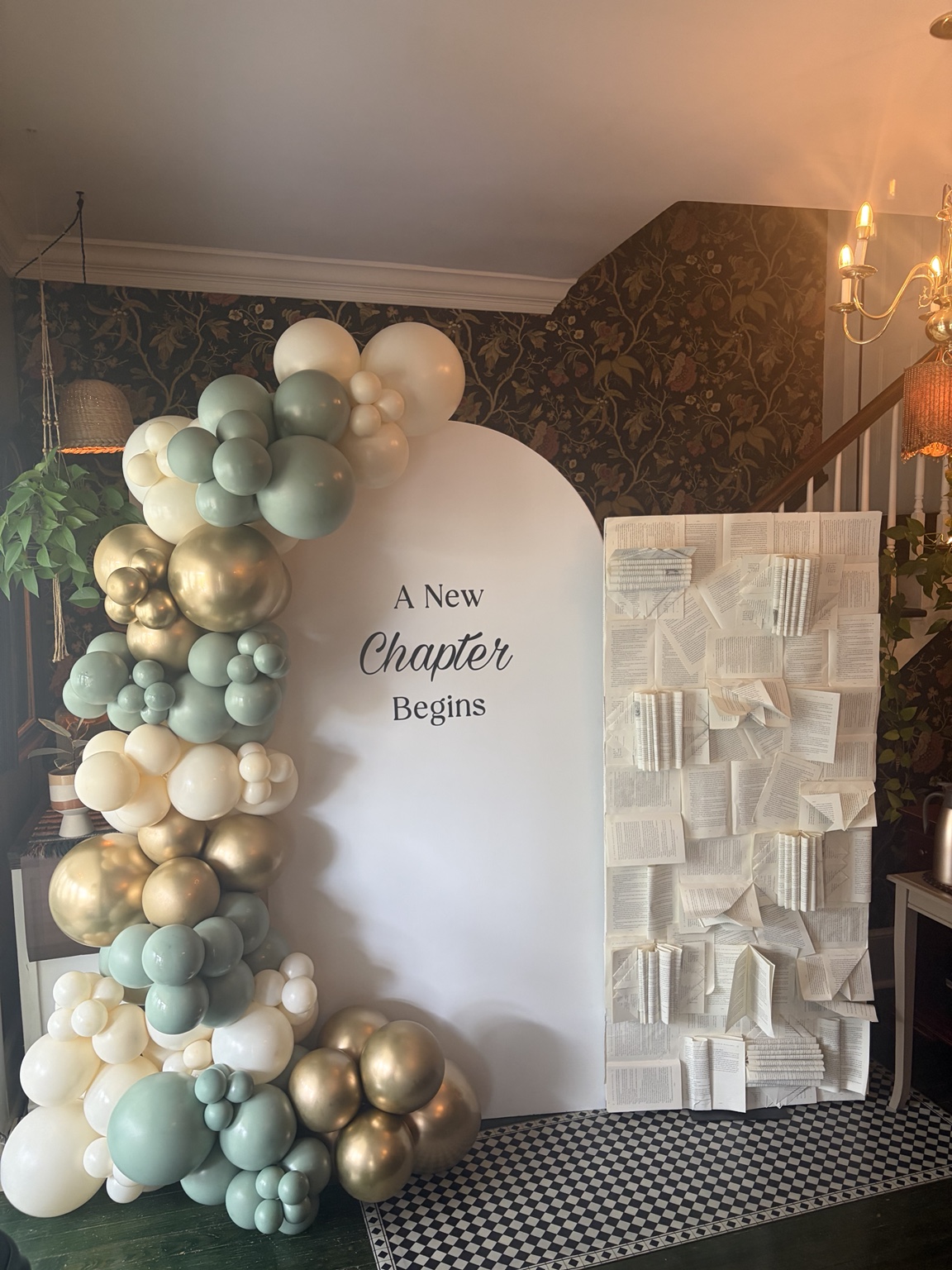 Sage, ivory and champagne balloons framing a New Chapter Begins backdrop beside an artful wall of book pages
