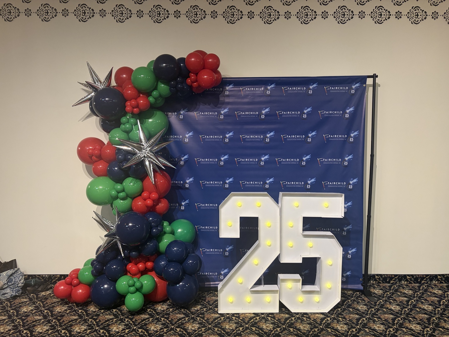 Red, green and navy branded balloon installation framing a Fairchild 25th-anniversary photo backdrop and lit marquee numbers