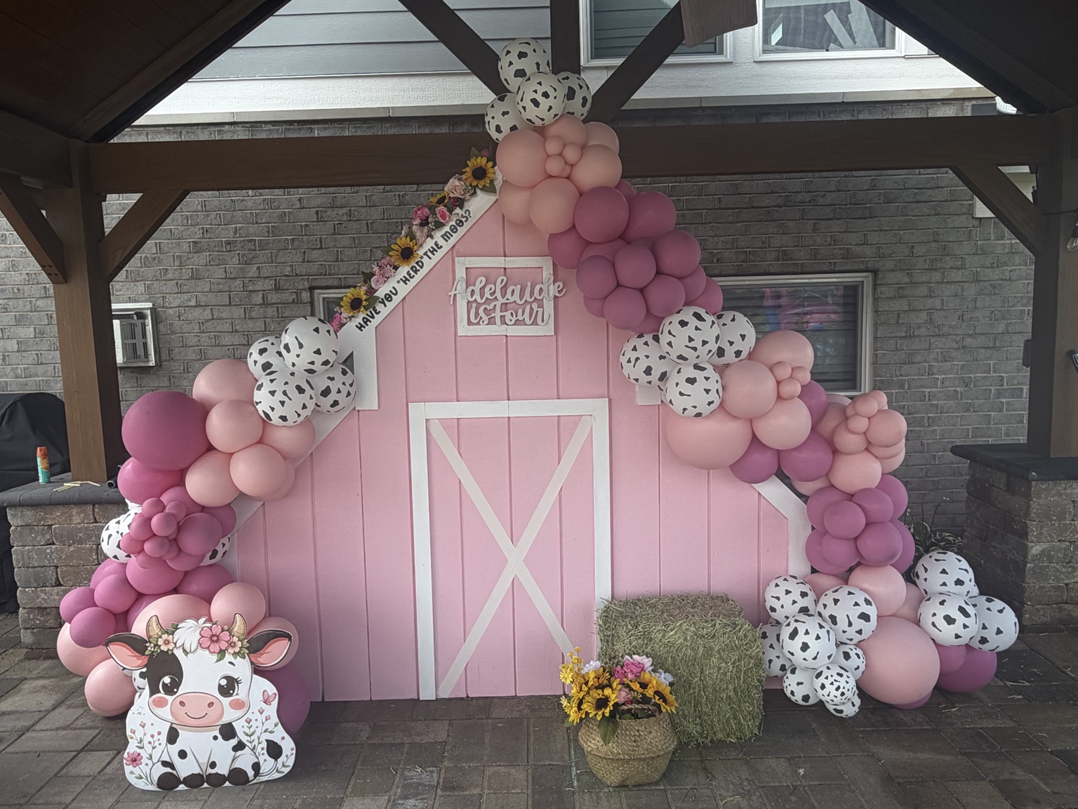 Pink barn backdrop framed by sculptural pink balloons, cow-print accents, sunflowers and farm-themed details