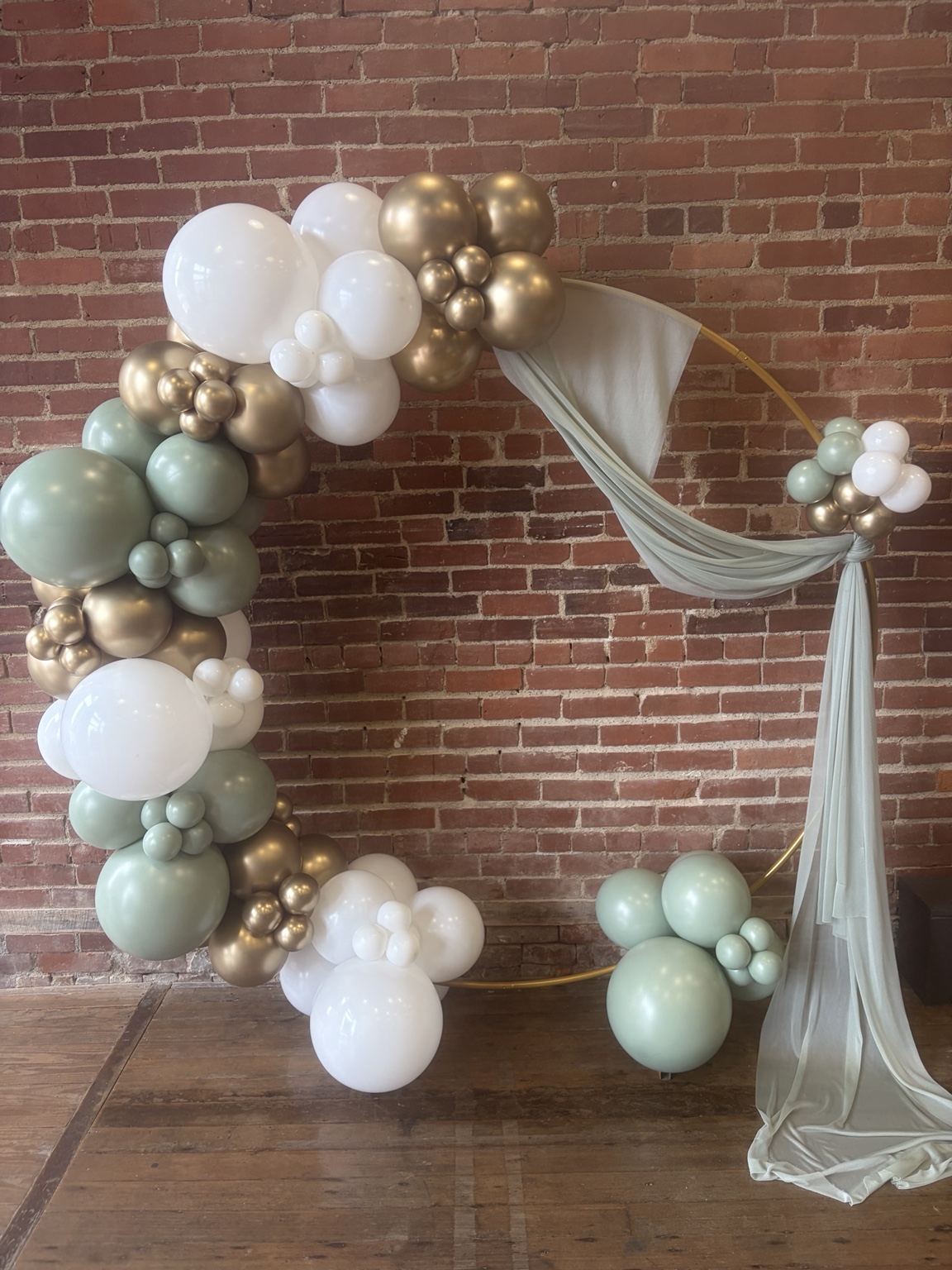 Sculptural sage, cream and champagne balloon installation framing a gold hoop with soft draped fabric against a brick wall