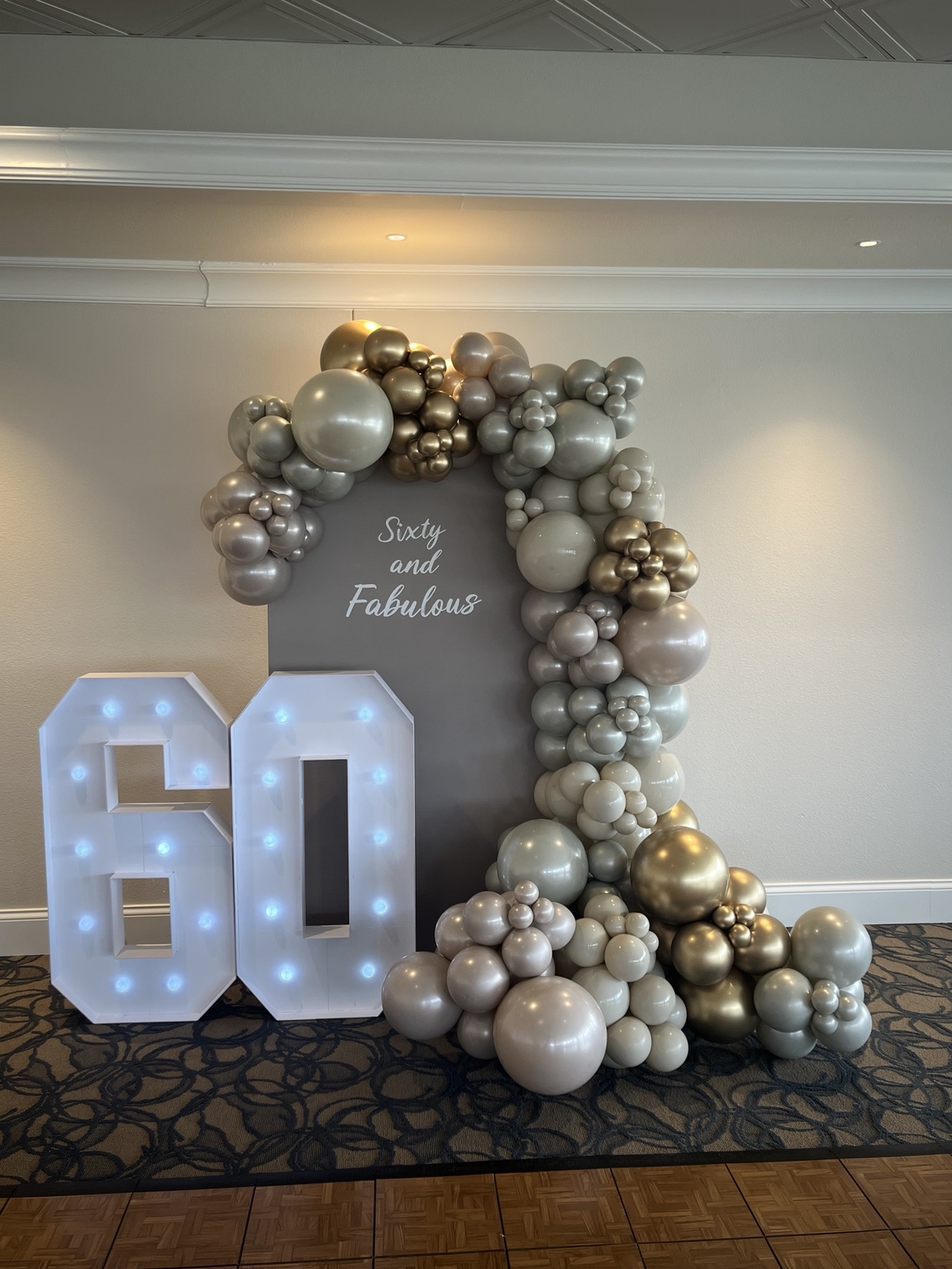 Layered champagne, silver and neutral metallic balloon arrangement alongside illuminated 60 marquee numbers